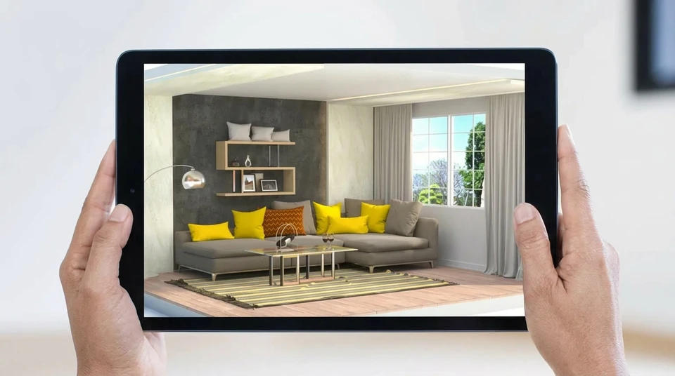 Personne tenant une tablette tactile affichant un canapé gris avec coussins orange en réalité augmentée dans un intérieur domestique minimaliste aux murs blancs et parquet bois clair